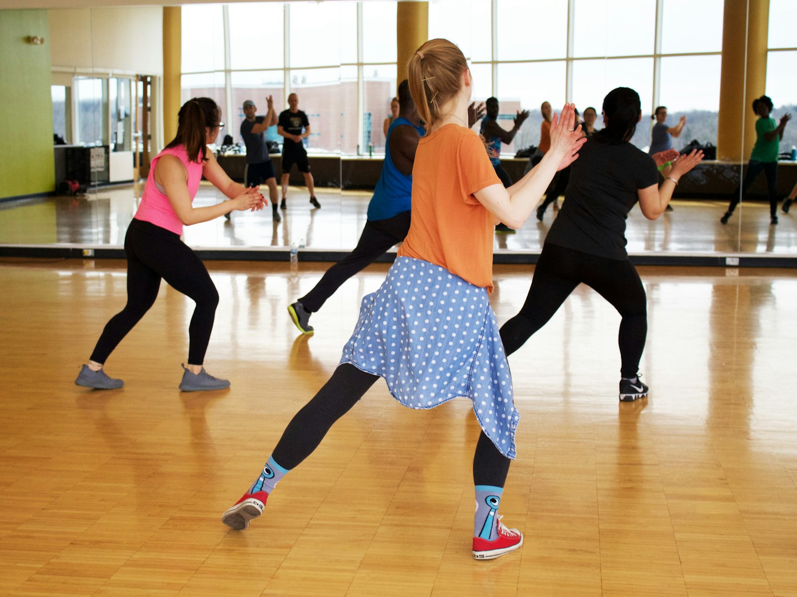A group dance class in motion in a bright studio with a mirror wall and wood floor.
